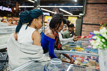 Group of african womans with shopping carts near refrigerator choose packs of dumplings in the supermarket.