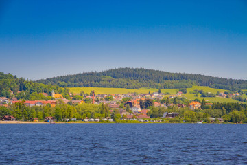 Fototapeta na wymiar The Lipno Dam - Horni Plana, Czech Republic, in a bright summer day. The lake is calm, has clean blue water. There are no clouds in the sky. Lot of trees on the background.