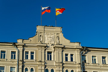 Fototapeta premium Close-up of top decoration of main facade Voronezh city administration building on Plekhanovskaya Street 10. Building was built in 1914-1915. Flags of Russia and region flying against blue sky