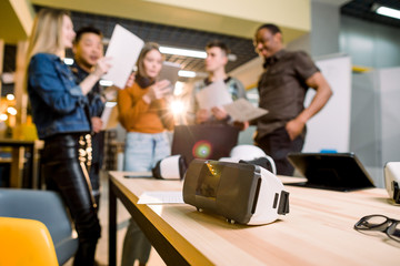 Fototapeta premium Blurred image of five multiethnical business persons are developing a project using modern gadgets and virtual reality goggles. Focus on the VR goggles on the table. technologies of the future