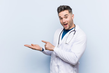 Young handsome doctor man excited holding a copy space on palm.