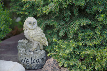 A garden owl figurine with a greeting standing on a large stone against a background of green fir...