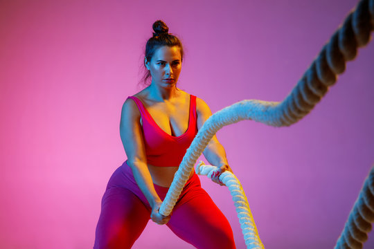 Young caucasian plus size female model's doing exercises on gradient purple background in neon light. Cross training with the rope. Concept of sport, healthy lifestyle, body positive, equality.