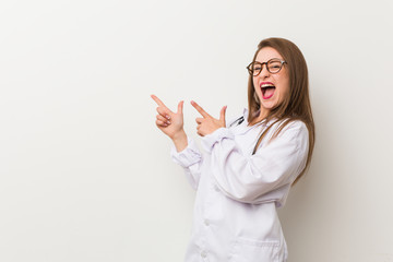 Young doctor woman against a white wall pointing with forefingers to a copy space, expressing...