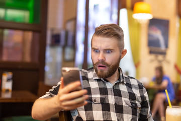 Shocked young hipster man drinking cocoa from a straw and looking at messages in social networks while sitting at a table in a cafe. The concept of chatting and social networks.