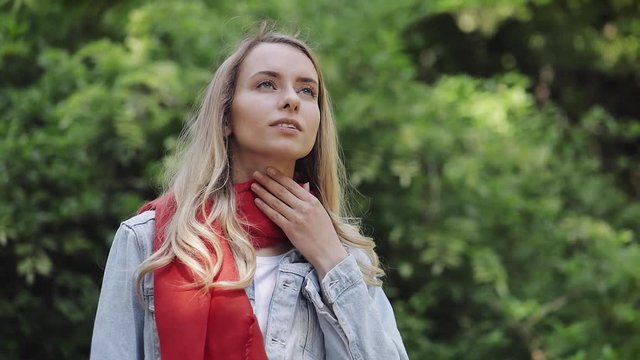 Sick young woman wearing red scarf feeling bad suffering from throat pain, sore throat, standing in the park. Painful Swallowing, health concept.