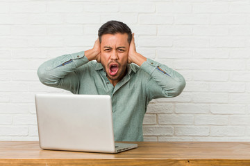 Young filipino man sitting working with his laptop covering ears with hands trying not to hear too loud sound.