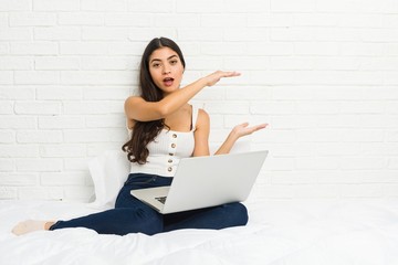 Young arab woman working with her laptop on the bed shocked and amazed holding a copy space between hands.