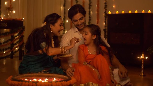 South Indian family dressed in traditional wear is celebrating Diwali at home - Happy Family. Family spending time together  eating sweets and celebrating Diwali - Festive colorful background with ...