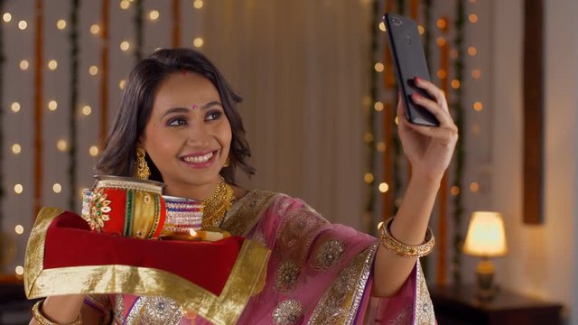 Beautiful Indian woman taking a selfie while waiting for her husband on the occasion of Karwa Chauth. Attractive young smiling wife taking a picture with puja thali in her hand - capturing memories...