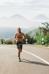 Man running topless in uphill on the asphalt road in hot summer weather. Panoramic mountain view on...