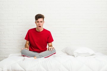 Young teenager student man on the bed screaming very angry and aggressive.
