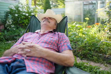 Senior hispanic man in hat sitting leaning back on chair sleeping in outdoor summer flower garden