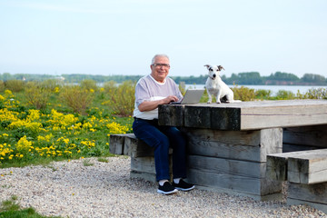 Obraz premium senior people, old age, lifestyle, distance learning concept. Smiling Senior man 70-75 years old sitting on wooden bench in summer park with dog jack russell terrier and working on laptop computer