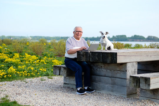 Senior People, Old Age, Lifestyle, Distance Learning Concept. Smiling Senior Man 70-75 Years Old Sitting On Wooden Bench In Summer Park With Dog Jack Russell Terrier And Working On Laptop Computer