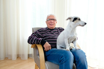 Happy Grandpa is sitting at chair in home with dog Jack Russell Terrier on his legs. Senior man 70-75 years old smiling at camera. Light window on background. Сoncept of happy old age.