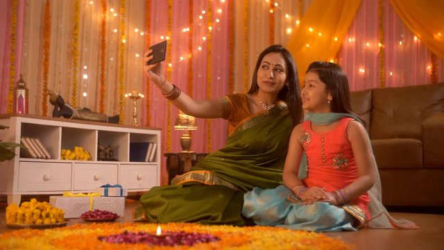 Festival occasion - young Indian mother is clicking a selfie with her daughter. Diwali decoration . Video footage of a happy mother and cute daughter wearing Indian festive dress. Using a smartphon...