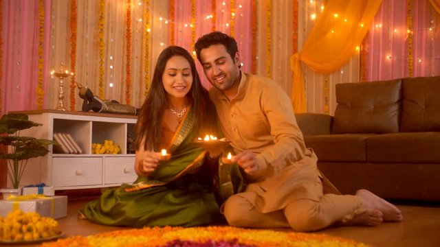 Diwali festival decoration - Young couple decorating rangoli with diyas. Colorful background with lights. Indian Stock Footage of young women in green saree and bangles decorating her house with he...