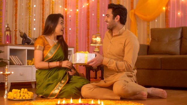 Diwali celebration - Handsome husband giving surprise gift to beautiful wife (Happy Indian Family). Indian Stock Footage of young couple wearing traditonal festive dress sitting on floor near rango...