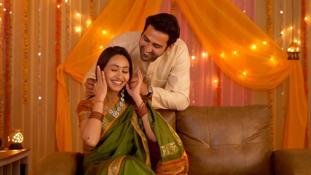 Indian Husband tying beautiful necklace around wife's neck as a gift - Couple in Love. Indian stock video of a lovable and caring husband-wife pair - Festival celebration at home. Colorful backgrou...
