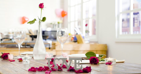 Festive table setting with love letter and flowers in vase glass for Valentine Day. roses petals around on wooden desk in dining room at home prepared for anniversary celebration concept lifestyle.