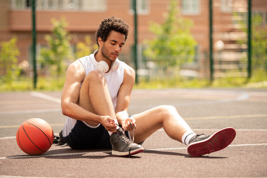 Young athlete sitting on basketball court and tying shoelace of sneaker