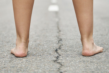 Rear view of bare feet of a young girl standing on the asphalt road close up. Dividing road lines...