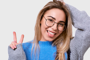 Closeup portrait of joyful young woman showing tongue and peace gesture while looking to the camera and standing against white background. Happy female wearing casual outfit, transparent glasses.