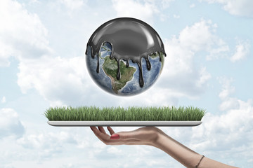 Female hand holding earth globe covered with black thick liquid on green grass model on blue sky background