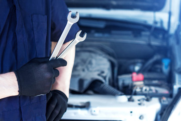 Repairman is repairing car at service station. Closeup mechanic hands in gloves are holding steel wrenches. Vehicle with open hood on background.  Modern auto repair shop with equipments and  tools.