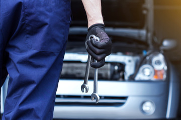 Repairman is repairing car at service station. Back view mechanic hands in gloves are holding steel wrenches. Vehicle with open hood on background.  Modern auto repair shop with equipments and tools.