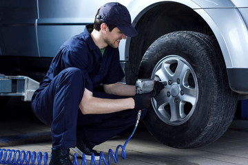 Smiling mechanic in blue jumpsuit is repairing car at service station garage. Repairman is removing wheel by electric wrench, tyre mounting equipment at workshop auto repair shop. Tire fitting concept