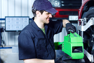 Smiling man mechanic is repairing red car at service station. Repairer in blue jumpsuit and cap is adjusting sensors on wheels. Process of wheels alignment camber check in workshop auto repair shop.