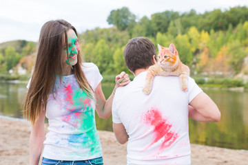 Obraz premium Festival holi, holidays, tourism, pet and nature concept - Portrait of woman and man with cat covered multi-colored dust