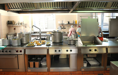 At the kitchen: gas-stoves, cooking utensils set on kitchen desk and cooks doing their job on a background