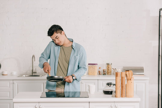 handsome asian man preparing breakfast on frying pan while talking on smartphone in spacious kitchen