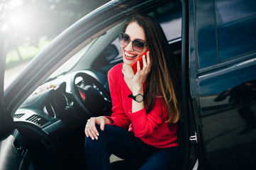 Fototapeta na wymiar Fashion cheerful woman calling on cell phone in her car. Stylish female model wearing sunglasses red sweater and trendy watch on hand speak with somebody on smartphone. Lifestyle People Travel concept