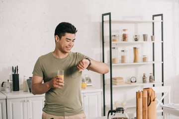 Fototapeta na wymiar cheerful asian man looking at watch while holding glass of orange juice