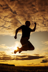 Silhouette of a young man in a jump against the backdrop of the setting sun and sky. The concept of happiness, activity, freedom, health. Man of dense build in a t-shirt and shorts. Place for text.