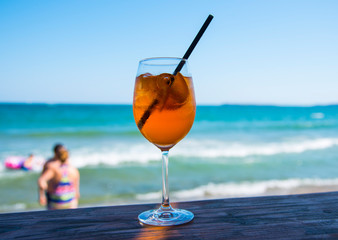 A glass with an orange cocktail an aperol with a black straw stands on a bar counter against the background of the sea and holidaymakers.