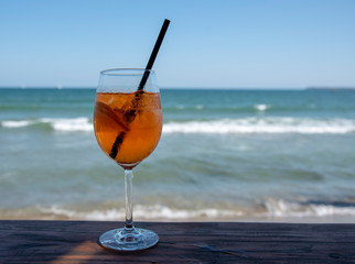 A glass with an orange cocktail an aperol with a black straw stands on a bar counter against the background of the sea and holidaymakers.