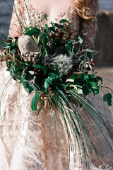 The bride is holding a bouquet of various exotic plantson a blurred background of the river and the city, selective focus