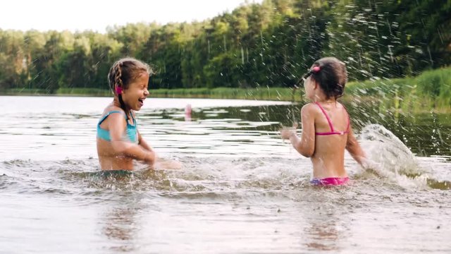 Two sisters in bathing suits splashing on the lake, near the pine trees in a picturesque place. Carefree having fun and enjoy life. 4k