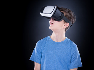 Happy teen boy wearing virtual reality goggles watching movies or playing video games, on black background. Cheerful teenager looking in VR glasses. Funny child experiencing 3D gadget technology.