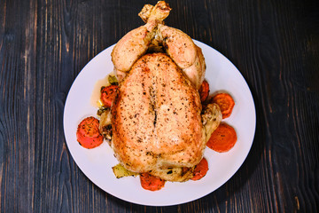 Festive dinner with baked bird and carrot. Fried chicken on black wood background, top view. Large whole grilled juicy chicken on a plate.
