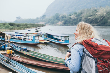 Fototapeta na wymiar Caucasian blonde woman with backpack stands on boat pier in Muang Ngoi village, Laos