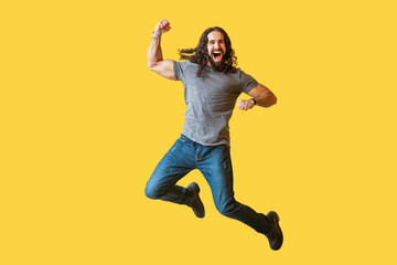 Fototapeta na wymiar Portrait of happy rejoicing bearded young man with long curly hair in casual grey tshirt jumping and celebrating his vivtory with amazed excited face. indoor studio shot isolated on yellow background.