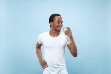 Half-length close up portrait of young african-american man in white shirt on blue background. Human emotions, facial expression, ad concept. Holding a glass and drinking water.