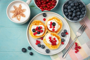 Fresh and tasty breakfast: cottage cheese pancakes with red currant and blueberry. Coffee in a cup with a pattern on the foam on a blue background. Close-up.