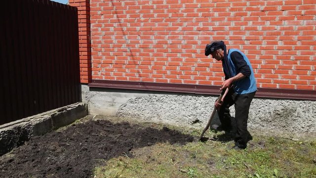 the older man, in a blue jacket, black pants, a rag on his head, digging up the ground with a shovel, near a high brick fence, in the afternoon in Sunny weather, slow shooting